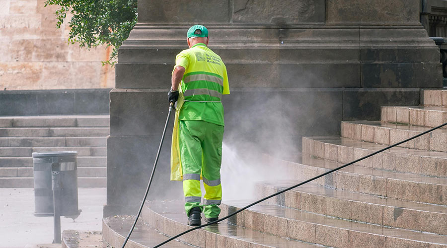 Worker in bright green uniform pressure washing outdoor stone steps, removing dirt and grime, with water spray and mist visible.